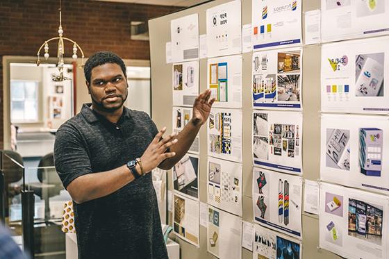Photo of a Chatham University design student presenting in front of a wall of printed design material. 