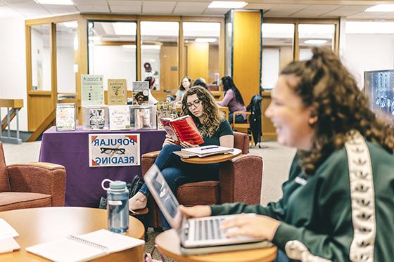 Photo of Chatham University students gathering in Jennie Mellon King Library, sitting in chairs and tables while working on laptops and reading books. 